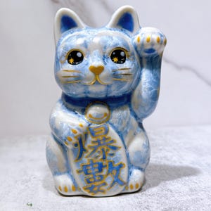 May include: A blue and white ceramic Maneki Neko cat figurine with a raised paw. The cat has gold accents on its eyes, nose, and collar. The figurine has Japanese characters on its chest.