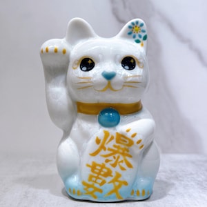 Può includere: Statuetta di gatto Maneki Neko in ceramica bianca con una zampa alzata. Il gatto ha occhi neri, naso blu e un collare giallo con un globo blu. La statuetta ha un motivo floreale sull'orecchio e testo dorato sulla pancia.