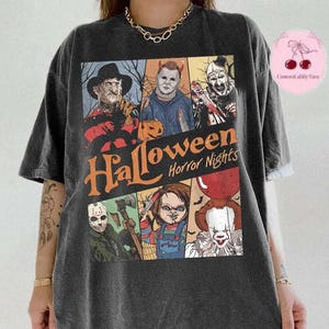 May include: A charcoal gray short-sleeve t-shirt featuring a graphic print of various horror movie characters. The text "Halloween Horror Nights" is printed in orange across the center. The design includes illustrations of Freddy, Michael, Art, Jason, Chucky, and Pennywise.