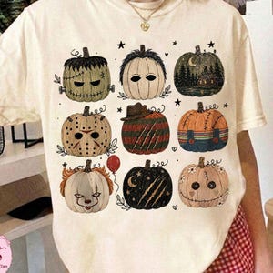 May include: A cream-colored short-sleeve t-shirt featuring a graphic print of nine stylized pumpkins. Each pumpkin is decorated to resemble a different horror movie character, including designs with stitches, masks, stripes, and dark forest silhouettes.