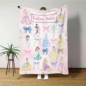Pu&ograve; includere: Coperta rosa con illustrazioni di principesse e fiocchi. La coperta ha uno sfondo rosa chiaro con vari personaggi di principesse nei loro abiti e fiocchi rosa e blu. Il testo "Custom Name" &egrave; visualizzato in alto.