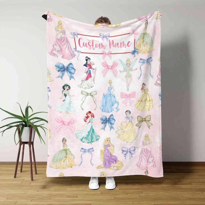 Pu&ograve; includere: Coperta rosa con illustrazioni di principesse e fiocchi. La coperta ha uno sfondo rosa chiaro con vari personaggi di principesse nei loro abiti e fiocchi rosa e blu. Il testo "Custom Name" &egrave; visualizzato in alto.