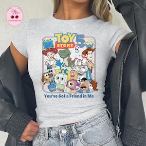 May include: A light gray t-shirt featuring a colorful graphic of characters from the animated film "Toy Story." The design includes the text "TOY STORY 5" and "You've Got a Friend in Me."
