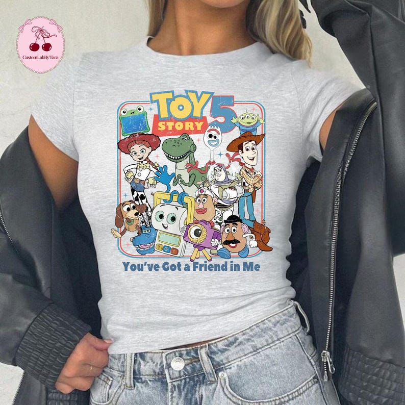 May include: A light gray t-shirt featuring a colorful graphic of characters from the animated film "Toy Story." The design includes the text "TOY STORY 5" and "You've Got a Friend in Me."