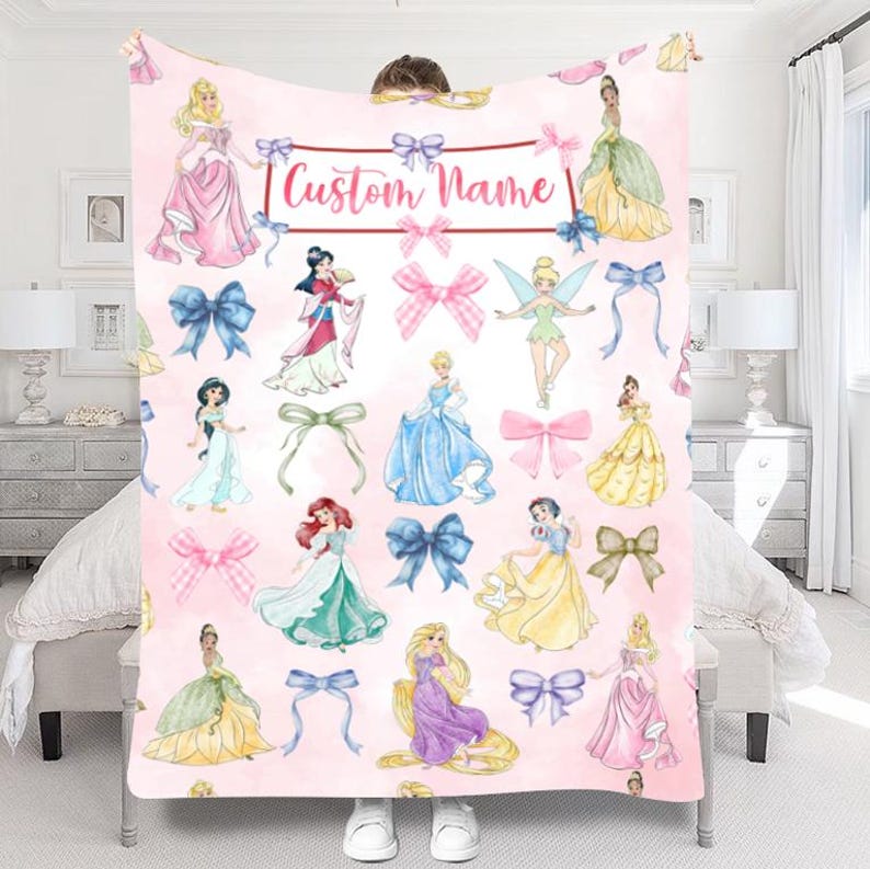 Pu&ograve; includere: Una coperta rosa con illustrazioni di principesse e fiocchi. La coperta ha il testo "Custom Name" in un banner. Le principesse indossano vari abiti e i fiocchi sono in tonalit&agrave; rosa e blu.