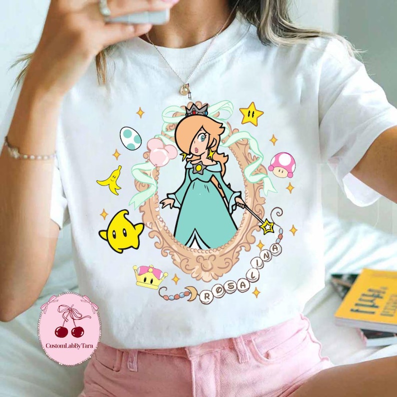 May include: White t-shirt featuring a cartoon illustration of Rosalina from the Super Mario franchise. The design includes a framed portrait of Rosalina, stars, a crown, and the word "Rosalina".