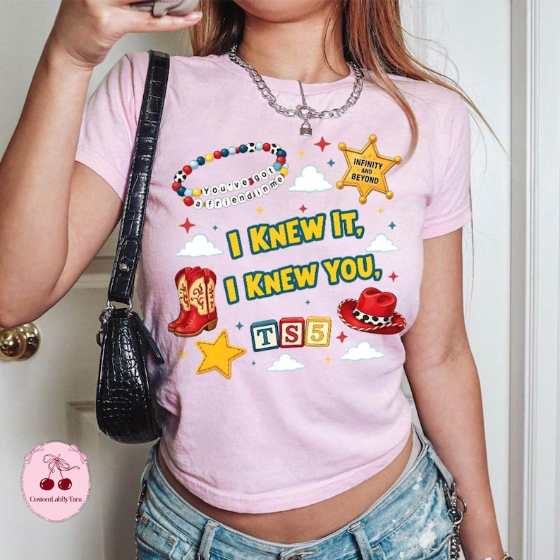 Op de afbeelding: Een lichtroze T-shirt met korte mouwen en een grafische print met een kralenarmband, een gouden sheriffster, rode cowboylaarzen, een rode cowboyhoed en alfabetblokken die TS5 spellen. De tekst luidt You've got a friend in me, Infinity and Beyond, I KNEW IT, I KNEW YOU.