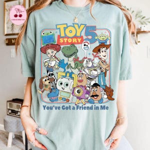 May include: A light blue t-shirt featuring a colorful graphic of characters from the movie "Toy Story". The design includes Woody, Buzz Lightyear, and other beloved characters, with the text "Toy Story 5" and "You've Got a Friend in Me".