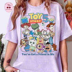 May include: A light purple t-shirt featuring a colorful graphic of characters from the movie "Toy Story 5". The design includes the text "TOY STORY 5" and "You've Got a Friend in Me".
