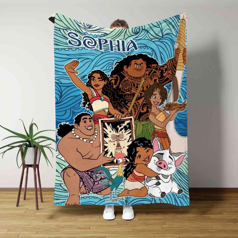 May include: A blue blanket featuring characters from the animated movie Moana, with the name "Sophia" in a stylized font. The blanket has a wave pattern background and cartoon illustrations of the characters.