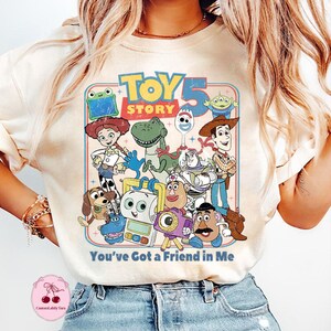 May include: Cream-colored t-shirt featuring a colorful graphic of Toy Story characters, including Woody, Buzz Lightyear, and Rex. The text "TOY STORY 5" is at the top, with "You've Got a Friend in Me" below. The design has a vintage, retro style.