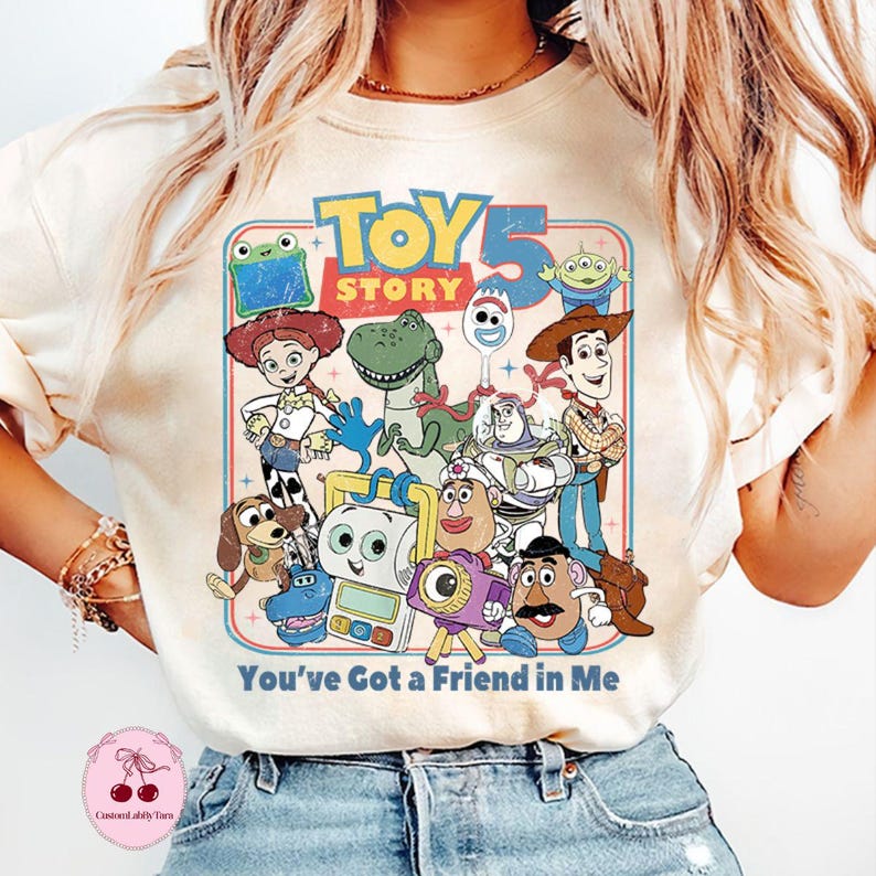 May include: Cream-colored t-shirt featuring a colorful graphic of Toy Story characters, including Woody, Buzz Lightyear, and Rex. The text "TOY STORY 5" is at the top, with "You've Got a Friend in Me" below. The design has a vintage, retro style.