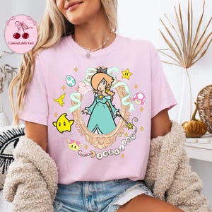 May include: A light pink t-shirt featuring a cartoon illustration of a character in a gold frame, with the name "ROSALINA" below. The design includes stars, a crown, and other game-related elements.