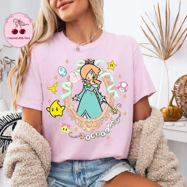 May include: A light pink t-shirt featuring a cartoon illustration of a character in a gold frame, with the name "ROSALINA" below. The design includes stars, a crown, and other game-related elements.