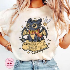 May include: Cream-colored t-shirt featuring a cartoon dragon wearing glasses and a scarf, sitting on books. A golden snitch and an owl are also depicted. The design includes stars and a wand, with the text "Customably Tara" at the bottom.