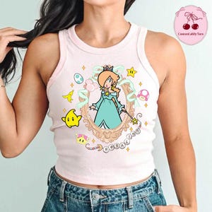 May include: A light pink tank top featuring a framed illustration of Rosalina from the Super Mario franchise. The design includes stars, a crown, and the name "Rosalina". The tank top is a casual, sleeveless garment.