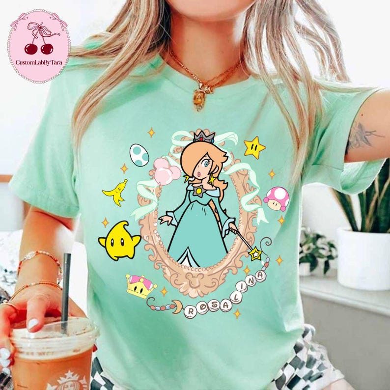 May include: A light green t-shirt featuring a cartoon illustration of Rosalina from the Super Mario franchise. The design includes a framed portrait of Rosalina, stars, a mushroom, and the word "Rosalina".