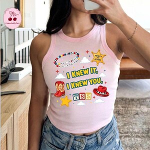 Op de afbeelding: Een lichtroze mouwloze tanktop met een grafische print van een kleurrijke kralenketting, een gele sheriffster, rode cowboylaarzen, een rode cowboyhoed en bouwblokken met de tekst TS5. De tekst op het shirt luidt I KNEW IT, I KNEW YOU.