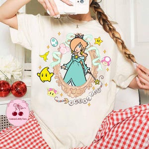 May include: Cream-colored t-shirt featuring a cartoon illustration of Rosalina from the Super Mario franchise. The design includes a framed portrait of Rosalina, surrounded by stars, a crown, and the word "Rosalina".