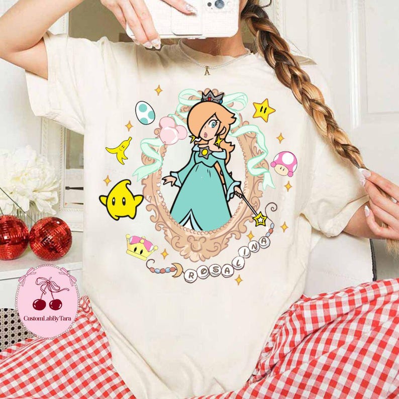 May include: Cream-colored t-shirt featuring a cartoon illustration of Rosalina from the Super Mario franchise. The design includes a framed portrait of Rosalina, surrounded by stars, a crown, and the word "Rosalina".