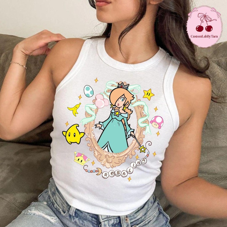 May include: White tank top featuring a cartoon illustration of Rosalina from the Super Mario franchise. The design includes a framed portrait of Rosalina, stars, and other game-related elements. The word "Rosalina" is written below the portrait.