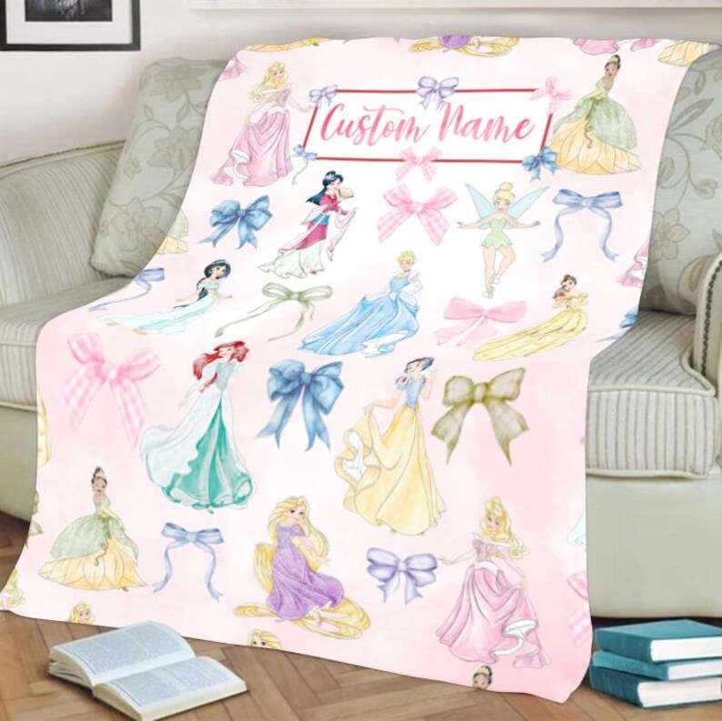 Pu&ograve; includere: Una coperta rosa con illustrazioni di principesse e fiocchi. La coperta ha uno sfondo rosa chiaro con varie principesse nei loro abiti e fiocchi sparsi sulla superficie. Il testo "Custom Name" &egrave; in un riquadro rosso.