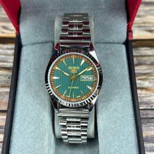 May include: A silver Seiko 5 automatic wristwatch with a green dial and a silver strap. The watch face shows the day and date, with gold hands and markers. The watch is in a grey box with a red interior.
