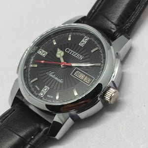 Op de afbeelding: Een Citizen automatisch polshorloge met een zwarte wijzerplaat en een zilverkleurige kast. Het horloge heeft een zwarte leren band en een datumweergave. De wijzerplaat heeft een radiaal patroon en de tekst "Citizen Automatic" is zichtbaar.