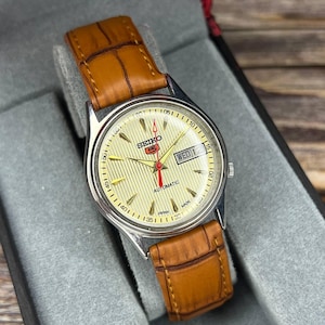 May include: A silver Seiko 5 automatic watch with a light yellow dial and a brown leather band. The watch face displays the day and date, with a red second hand. The watch is presented in a gray box.