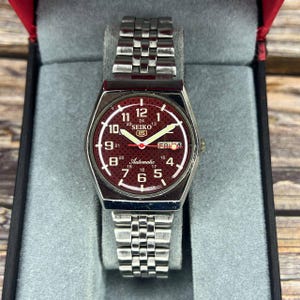 May include: A Seiko automatic wristwatch with a burgundy dial and a silver-tone stainless steel strap. The watch face features white numerals, hands, and the day of the week. The watch is displayed in a grey box with a red interior.