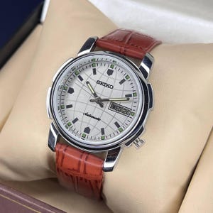 May include: A Seiko automatic wristwatch featuring a silver dial with a globe grid pattern, silver hands, and green accents. The watch has a day-date window at the three o'clock position and a brown leather strap with a textured crocodile-style finish.