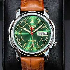May include: A Seiko automatic wristwatch with a green dial and gold-tone hands and markers. The watch features a brown leather strap and a stainless steel case. The day is displayed in Spanish. The watch is displayed on a black surface.