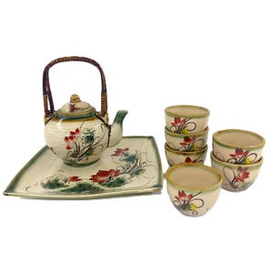 Puede incluir: Un juego de té de cerámica que incluye una tetera con asa trenzada, una bandeja cuadrada y cuatro tazas pequeñas. Cada pieza es de color blanco roto con flores de loto rojas, hojas verdes y un borde verde oscuro pintados a mano.