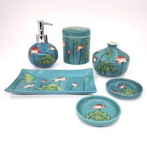 Puede incluir: Un juego de baño de cerámica turquesa con un diseño de flor de loto. El juego incluye un dispensador de jabón, un tarro con tapa, un jarrón decorativo, una bandeja rectangular y dos platos redondos pequeños. Las flores de loto son rojas y blancas.