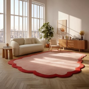 May include: A large, pink rug with a scalloped edge and a red border sits on a hardwood floor. The rug is in a living room with a cream-colored sofa, wooden furniture, and large windows overlooking a cityscape.