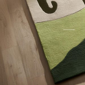 May include: A section of a green, beige, and dark green rug with an abstract design. The rug has a textured surface and is placed on a light brown wooden floor. The text "ChhayaCarpetsDesign" is visible on the rug.