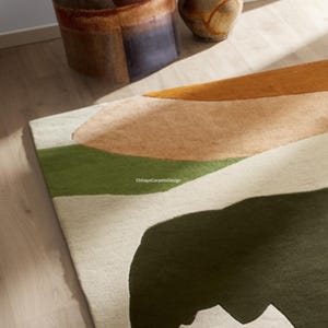 May include: A modern area rug with an abstract design. The rug features a cream-colored background with large, organic shapes in olive green, tan, and mustard yellow. The rug is made of a soft, textured material.