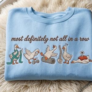 May include: A light blue crewneck sweatshirt featuring an embroidered graphic of five white geese in various poses with the text "most definitely not all in a row" in brown script. The geese are shown exercising, stretching, and resting.