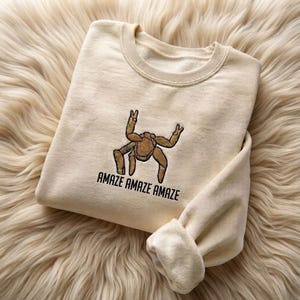 May include: A cream-colored crewneck sweatshirt featuring an embroidered brown tarantula graphic centered on the chest. Below the spider, the text "AMAZE AMAZE AMAZE" is embroidered in black capital letters. The garment has long sleeves and a ribbed collar.