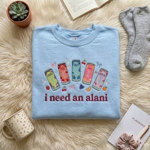 May include: Light blue sweatshirt with embroidered colorful beverage cans and the phrase "i need an alani" in maroon. The cans feature fruit designs and the sweatshirt is on a fluffy, white surface.