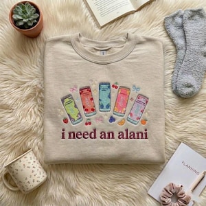May include: Beige crewneck sweatshirt with embroidered colorful beverage cans and the phrase "i need an alani" in maroon. The sweatshirt features a round neckline and long sleeves. The design includes fruit and bow accents.
