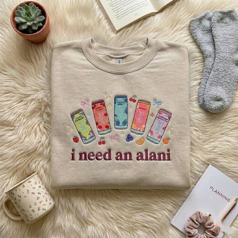 May include: Beige crewneck sweatshirt with embroidered colorful beverage cans and the phrase "i need an alani" in maroon. The sweatshirt features a round neckline and long sleeves. The design includes fruit and bow accents.