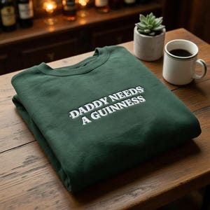 May include: A folded, dark green sweatshirt with the text "DADDY NEEDS A GUINNESS" embroidered in white. The sweatshirt is on a wooden surface, with a coffee mug and a small potted plant in the background.