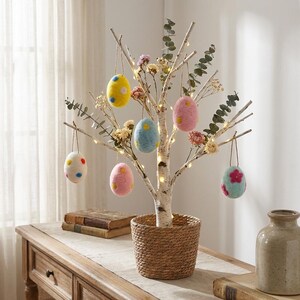 May include: A decorative Easter tree with birch branches, adorned with colorful felt egg ornaments. The eggs are in shades of yellow, pink, and blue, with floral and polka dot patterns. The tree sits in a woven basket, with fairy lights.