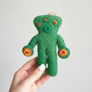 Könnte beinhalten: Handgefertigte grüne Alien-Puppe aus Filz mit drei roten Augen und orangefarbenen und gelben Akzenten an den Händen. Die Puppe hat eine rot-weiße Schnur zum Aufhängen. Die Puppe wird in einer Hand gehalten.