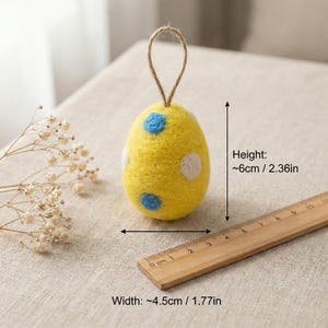 May include: A yellow felt Easter egg ornament with blue and white polka dots. The egg is approximately 2.36 inches tall and 1.77 inches wide, with a jute loop for hanging. A wooden ruler is next to the egg.
