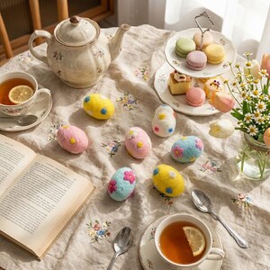 May include: A still life featuring a floral-patterned teapot, teacups with lemon slices, and decorative Easter eggs. The eggs are in various colors, including pink, yellow, and blue, with floral and polka dot designs. A two-tiered dessert stand holds macarons and pastries.