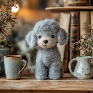 Könnte beinhalten: Eine handgefertigte graue Pudel-Hundefigur mit flauschiger Textur. Der Hund hat ein hellbeige Gesicht und dunkle Augen. Er sitzt auf einem Holzregal, mit einer Tasse und einem dekorativen Krug in der Nähe. Der Hintergrund umfasst Bücher und getrocknete Pflanzen.