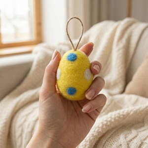 May include: A yellow felt Easter egg ornament with white and blue polka dots. The egg has a loop of twine for hanging. The ornament is held in a hand.