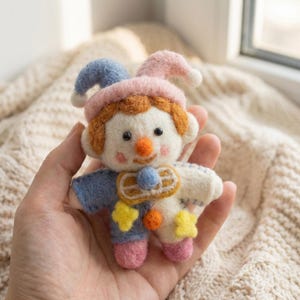 May include: A handmade felt clown doll with a blue and pink hat, orange hair, and a round orange nose. The doll has a blue shirt, white body, and pink feet. Yellow star and orange circle accents are sewn on the doll.